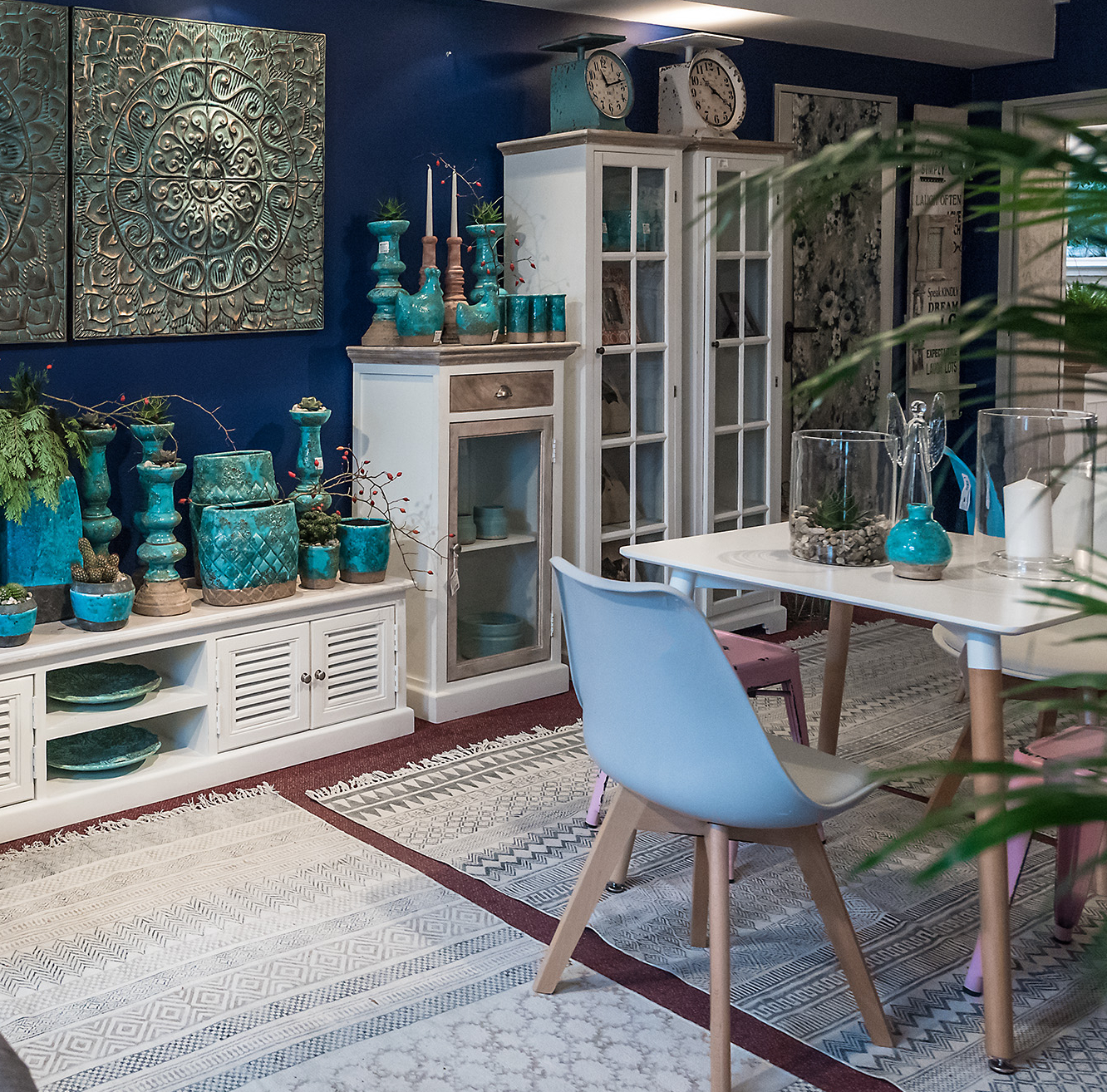 Salon z meblami Belldeco Bristol White – białe witryny i komody z drewnianymi elementami, zestawione z turkusowymi dekoracjami i nowoczesnym stołem jadalnianym.