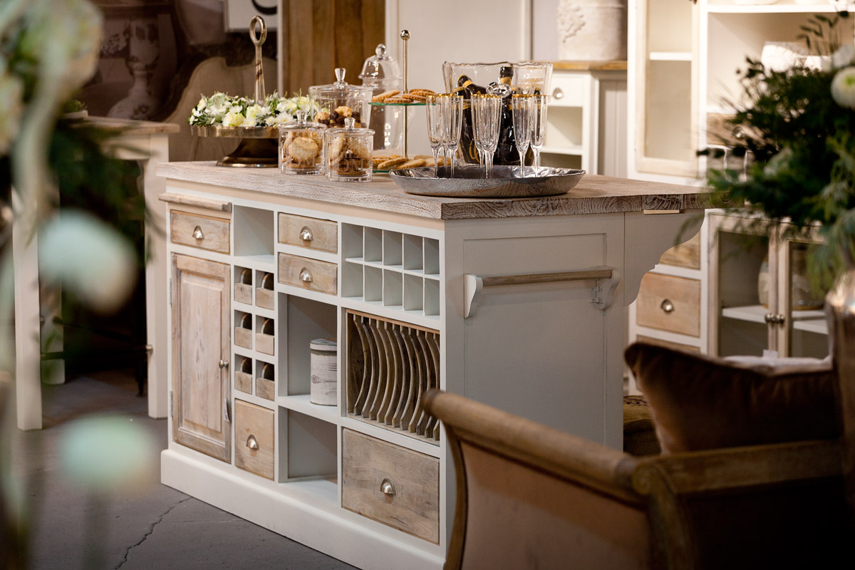Wyspa kuchenna Bristol White Belldeco w stylu prowansalskim z drewnianym blatem, szufladami i półkami na wino oraz talerze, udekorowana szklanymi pojemnikami ze słodkościami i kieliszkami.