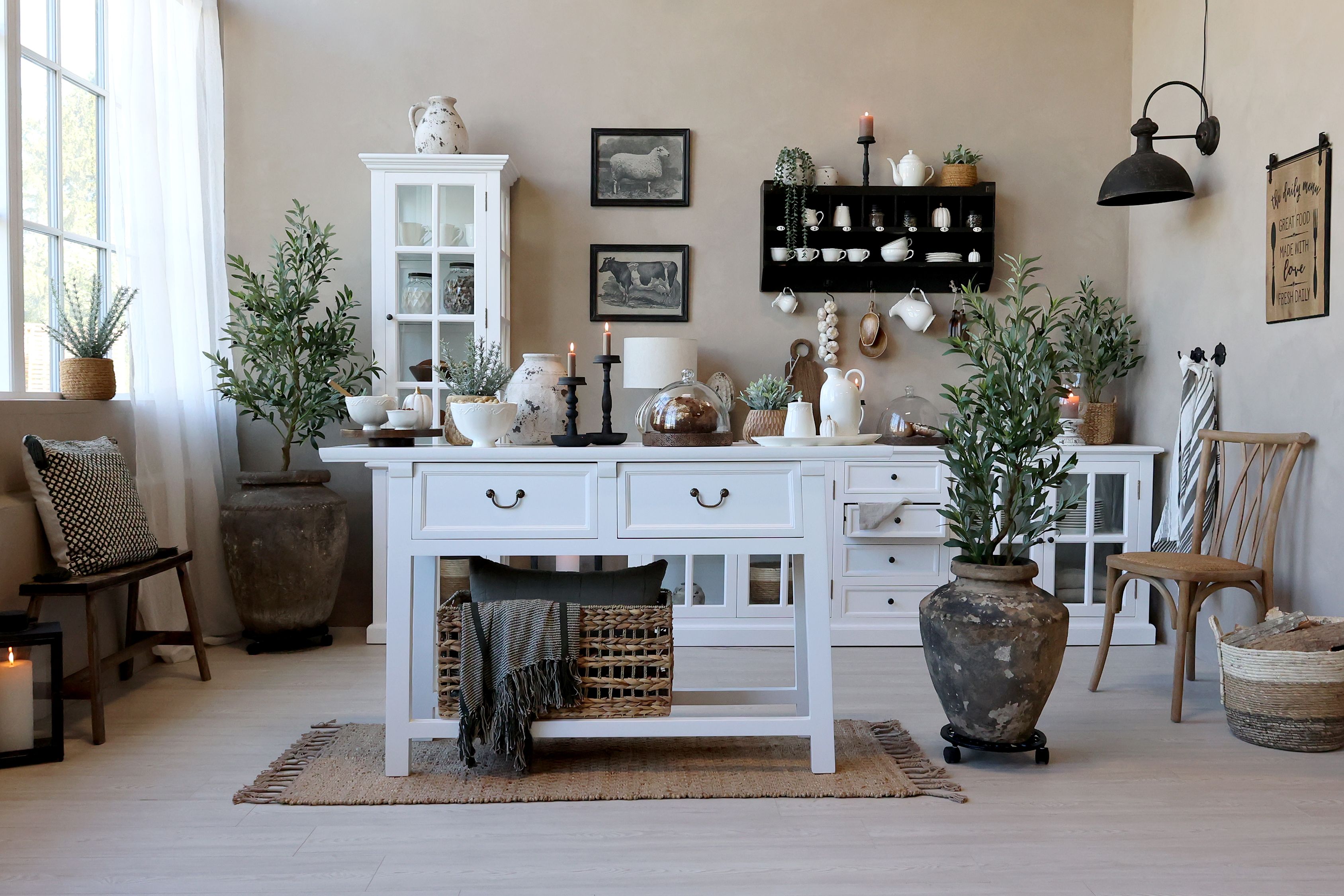 Stylowa kuchnia w klimacie prowansalskim urządzona białymi meblami z witrynami i szufladami, drewnianym stołem i naturalnymi dodatkami. Duże donice z zielonymi roślinami, kosze wiklinowe oraz ceramiczne naczynia tworzą przytulne i harmonijne wnętrze pełne światła i elegancji.