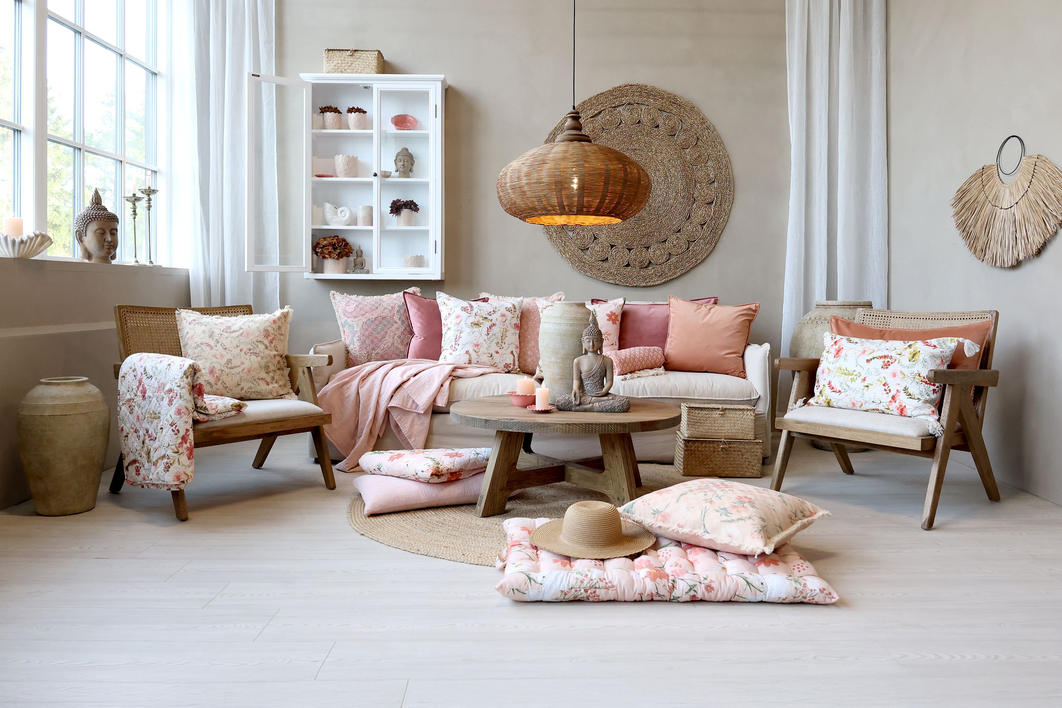 Przytulny salon w stylu boho z sofą i fotelami z drewnianymi elementami, udekorowany kolorowymi poduszkami i pledami w pastelowych odcieniach różu. Wnętrze uzupełniają naturalne dodatki z rattanu, wikliny i lnu, okrągły stolik kawowy oraz dekoracje inspirowane naturą i orientem.