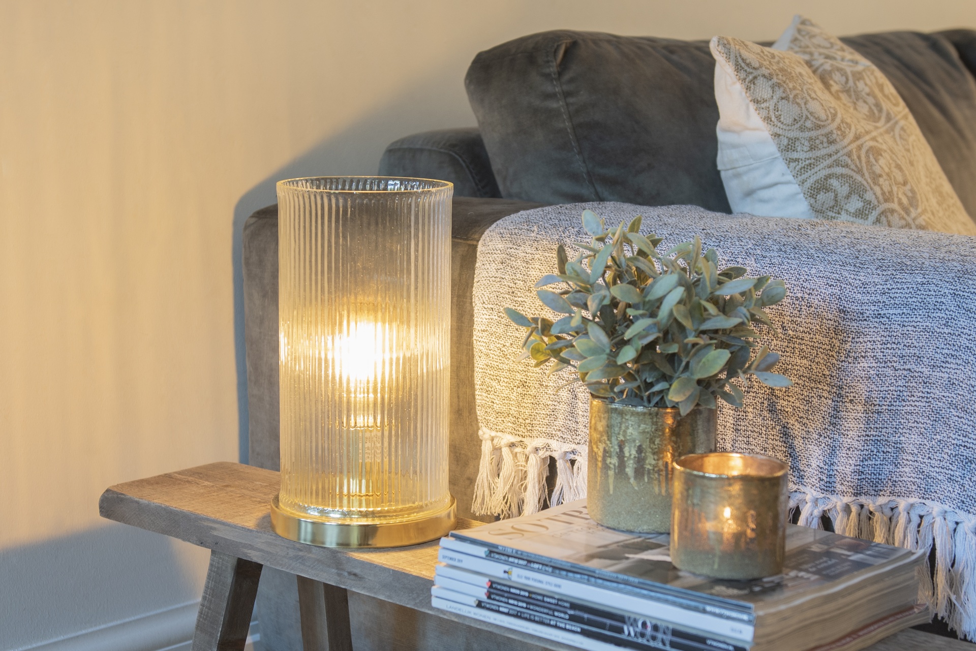 Przytulny kącik z lampą stołową o złotych akcentach, świecami i doniczką z rośliną ustawionymi na drewnianym stoliku obok sofy. Odpowiednio dobrane dodatki – miękki pled, dekoracyjne poduszki i ciepłe światło – sprawiają, że wnętrze staje się bardziej nastrojowe i przyjazne.