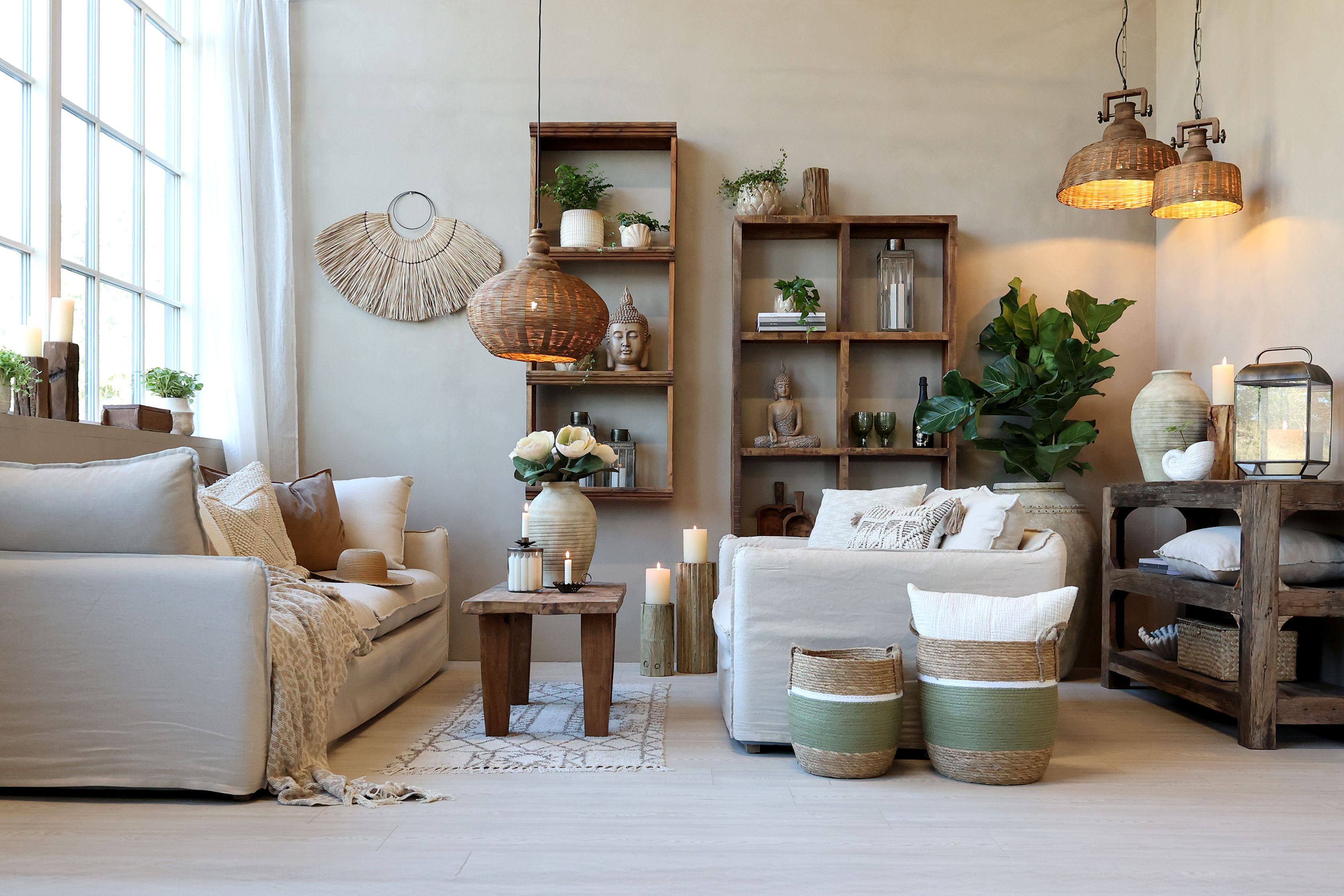 Przytulny salon w naturalnym stylu z sofami w jasnych barwach, wiklinowymi lampami i świecami, których ciepłe światło ociepla wnętrze i tworzy relaksującą atmosferę. Drewniane meble i zielone rośliny dopełniają harmonijną aranżację.