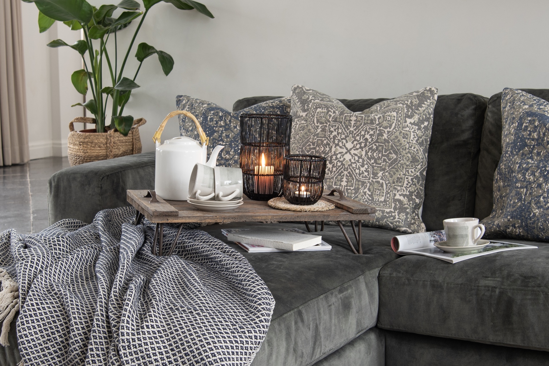 Przytulny salon z ciemnoszarą sofą udekorowaną wzorzystymi poduszkami i pledem. Na drewnianej tacy stoją kubki, dzbanek oraz świeczniki z zapalonymi świecami, które dodają wnętrzu ciepła i nastroju. Roślina w plecionym koszu uzupełnia naturalny charakter aranżacji.