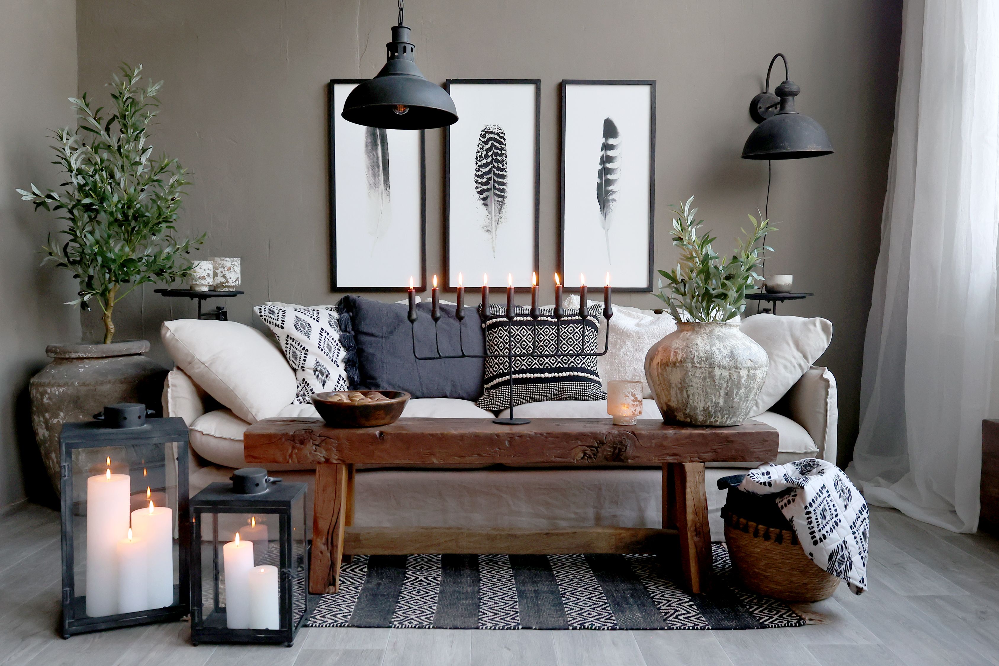 Przytulne wnętrze salonu z beżową sofą pełną poduszek, drewnianym stolikiem, dużymi latarniami ze świecami i dekoracyjnymi roślinami. Naturalne materiały, ciepłe światło i stonowane barwy tworzą harmonijną przestrzeń idealną do wypoczynku.