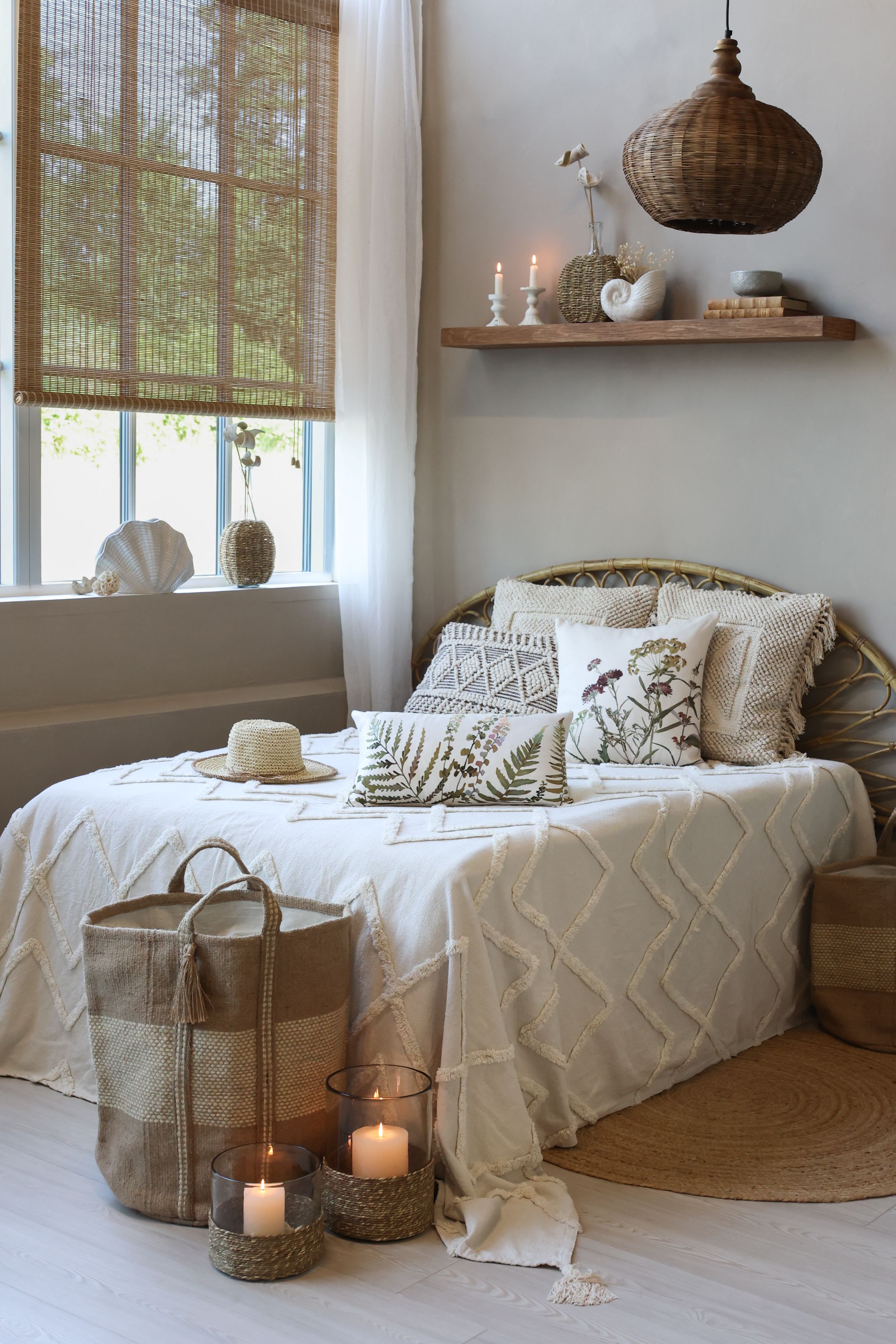 Sypialnia boho z rattanowym wezgłowiem, poduszkami w botaniczne wzory i naturalnymi dodatkami – kosze, świece i wiklinowa lampa.